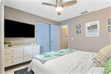 Coastal-inspired bedroom with ceiling fan, wall-mounted flat-screen TV above a white dresser, light-blue louvered closet doors, and a bed topped with a green tropical leaf throw.