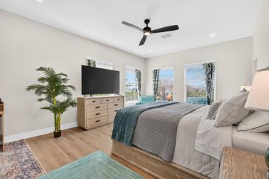 Bright coastal-style master bedroom with king bed and gray bedding, wood dresser with flat-screen TV, ceiling fan, turquoise chairs, potted palm and large sunny windows