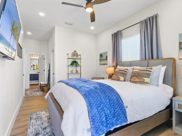 Bright modern coastal master bedroom interior with king bed, gray upholstered headboard, white linens and textured blue throw, patterned accent pillows, wood floors, ceiling fan, gray curtains, wall-mounted TV showing a beach scene, decorative shelving and open doorway to an en-suite bathroom.