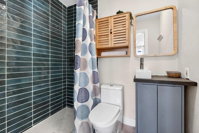 Cozy modern bathroom with dark green subway-tile shower, circular-patterned shower curtain, hexagon floor tile, white one-piece toilet, wooden wall cabinet, square rattan mirror, and gray chevron vanity