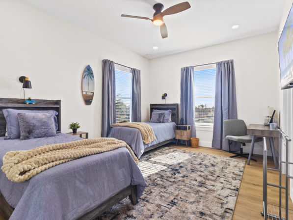 Sunny coastal-style twin bedroom with two gray-blue beds, chunky beige knit throws, wooden headboards, textured area rug, ceiling fan, desk and windows with navy curtains letting in bright daylight.