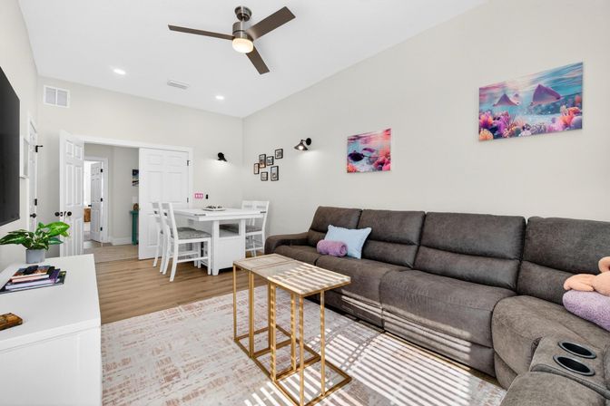 Sunlit modern living room interior with a large gray sectional sofa, pastel ocean-themed wall art, gold nesting coffee tables, white TV console and a compact white dining nook on warm hardwood floors under a ceiling fan.