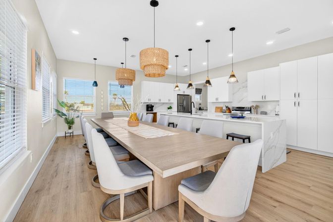 Bright, airy open-concept kitchen and dining area with a long light-wood table seating ten, upholstered chairs, white marble-topped island and cabinets, woven rattan and black pendant lights, large windows with blinds, potted plant, and light oak floors.
