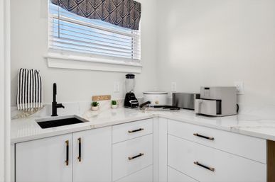 Bright modern white L-shaped kitchen with marble-look quartz countertops, black sink and faucet, striped hand towel and patterned window valance; counter holds a blender, slow cooker and stainless air fryer above white cabinets with black-and-brass handles.