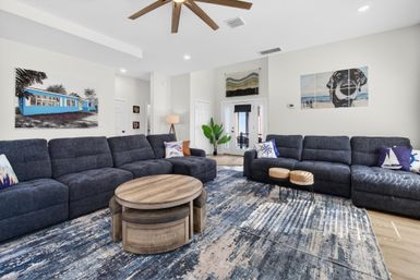 Spacious coastal-inspired living room with two dark gray sectional sofas, nautical throw pillows, round wooden nested coffee tables on a blue abstract area rug, ceiling fan, potted plant, and coastal-themed wall art.