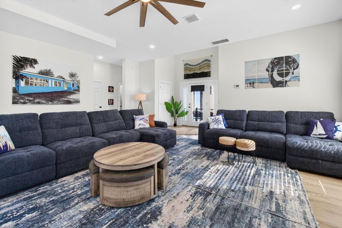Spacious coastal-inspired living room with two dark gray sectional sofas, nautical throw pillows, round wooden nested coffee tables on a blue abstract area rug, ceiling fan, potted plant, and coastal-themed wall art.