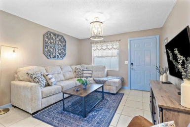 Cozy bright living room with beige L-shaped sectional, patterned throw pillows, navy blue area rug, wood coffee table with plant, TV on a wood console, decorative wall art and a light-blue front door.