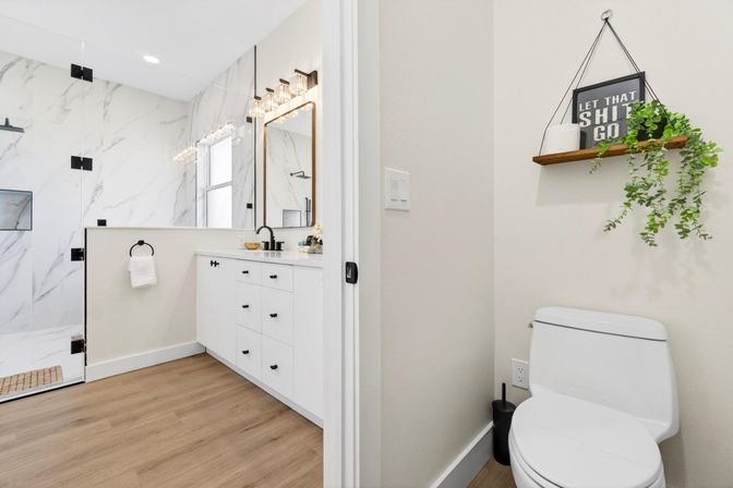 Bright modern bathroom with white vanity and black fixtures, glass walk-in shower with marble tile, wood-look floor, toilet beneath a hanging shelf with trailing plant and framed sign reading 'Let that shit go'.