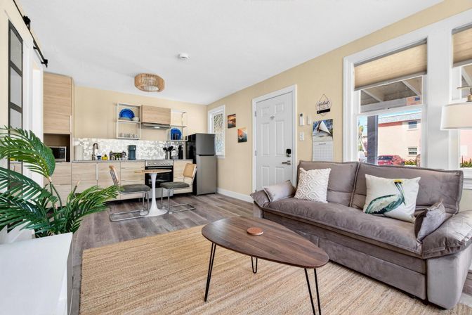 Bright, airy open-plan living room and kitchenette in a modern studio apartment — gray sofa with bird pillow, wood coffee table, jute rug, light wood cabinets, stainless fridge, small dining nook, large windows and indoor plant.