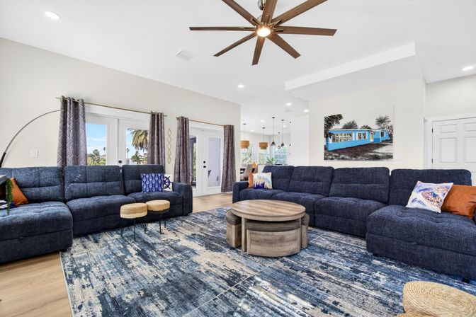 Spacious modern open-concept living room with navy sectional sofas, round wooden coffee tables, textured blue area rug, large ceiling fan, patterned curtains and glass doors showing palm trees outside