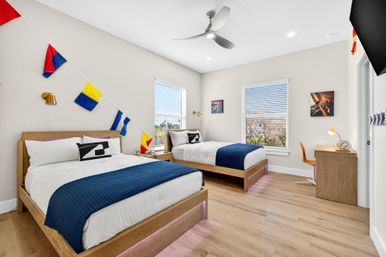 Bright modern coastal-style shared bedroom with two wooden platform beds, navy throws and playful nautical signal-flag bunting, light wood floors, study desk, windows with blinds and ceiling fan.