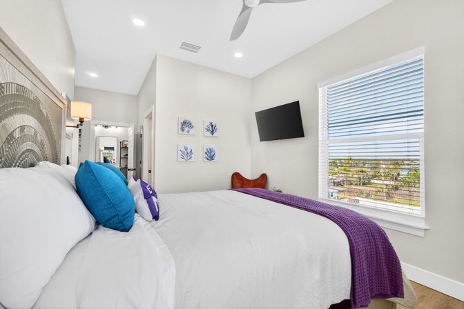 Bright coastal-inspired bedroom with white linens, blue accent pillows, purple throw, wall-mounted TV and palm-tree neighborhood view through blinds.