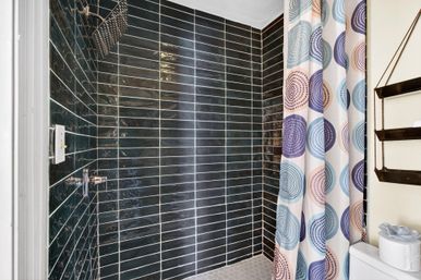Modern bathroom shower with glossy black subway tiles, hexagon floor tile, chrome rain showerhead and playful pastel circular-pattern shower curtain beside toilet and wall shelves.
