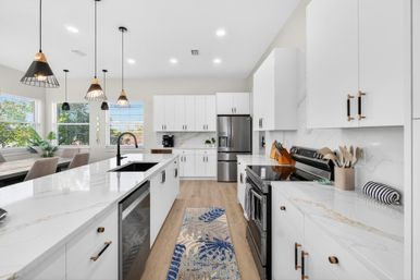 Bright modern open-concept white kitchen with marble countertops and large island, black faucet, pendant lights, stainless steel refrigerator and stove, hardwood floors, dining nook by sunny windows and a patterned runner rug.