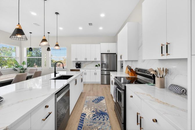 Bright modern open-concept white kitchen with marble countertops and large island, black faucet, pendant lights, stainless steel refrigerator and stove, hardwood floors, dining nook by sunny windows and a patterned runner rug.
