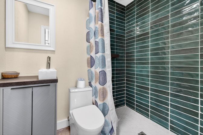 Modern small bathroom featuring a deep green horizontal subway-tile walk-in shower, white hex-tile floor, white toilet, gray vanity with vessel sink, and a colorful circular-pattern shower curtain.