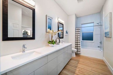 Bright modern residential bathroom with a long white double-sink vanity, black-framed mirrors, orchid centerpiece, wood-look floor, and a tub/shower with striped curtain and frosted window.