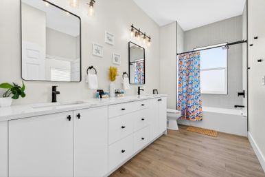 Bright modern bathroom featuring a white double vanity with black hardware and faucets, two rectangular mirrors with Edison-style lights, wood-look flooring, and a tub with a colorful blue-and-orange floral shower curtain and window.