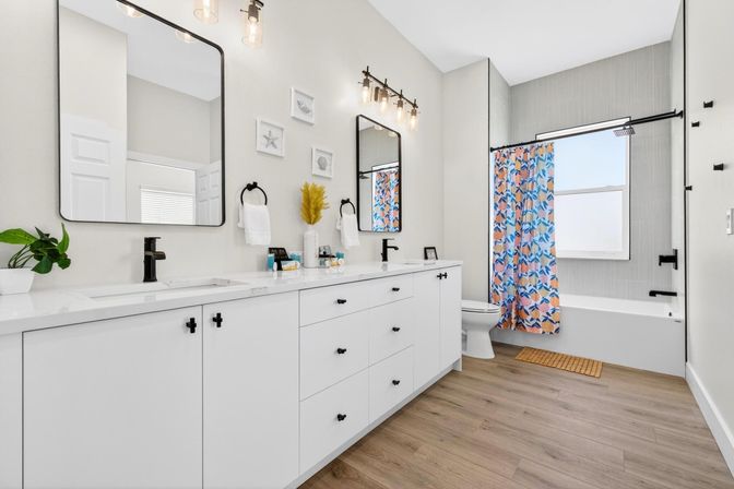 Bright modern bathroom featuring a white double vanity with black hardware and faucets, two rectangular mirrors with Edison-style lights, wood-look flooring, and a tub with a colorful blue-and-orange floral shower curtain and window.