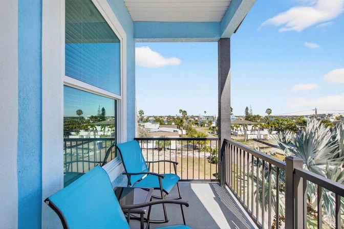 Sunny coastal balcony with bright turquoise patio chairs, black metal railing, palm trees and distant water view under a clear blue sky