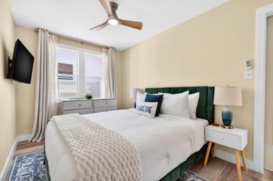 Sunny cozy bedroom with green velvet headboard, white bedding and chunky knit throw, ceiling fan, wall-mounted TV, bedside lamp and dresser under double window.