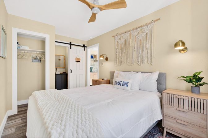 Cozy boho bedroom with fluffy white comforter, macrame wall hanging, wooden nightstands, brass wall sconces, potted plant, sliding barn door closet, and modern wooden ceiling fan