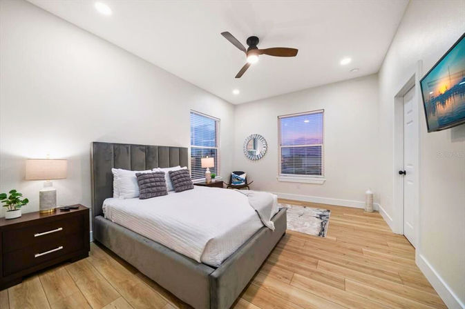 Modern master bedroom with upholstered grey bed, wood-look floors, ceiling fan, nightstands with lamps, twin windows showing a sunset and wall-mounted TV.
