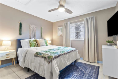 Bright coastal-style bedroom with queen bed, tropical leaf bedding, macramé wall hangings, ceiling fan, blue area rug and bedside tables
