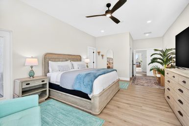 Bright coastal-style master bedroom with king bed, light wood furniture, teal accents, ceiling fan, hardwood floors, large TV and potted palm