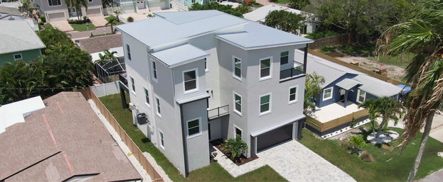 Aerial view of a modern three-story coastal home with light-gray stucco and metal roof, paver driveway, small lawn, palm trees and neighboring houses in a sunny beach neighborhood.