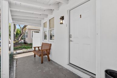 Sunlit covered front porch entryway of a small white house with a wooden bench, white paneled door labeled “1”, wall lantern, and a small landscaped yard visible beyond.