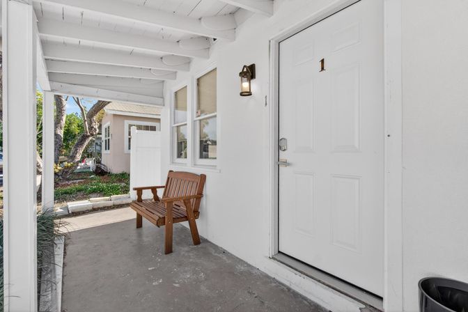 Sunlit covered front porch entryway of a small white house with a wooden bench, white paneled door labeled “1”, wall lantern, and a small landscaped yard visible beyond.