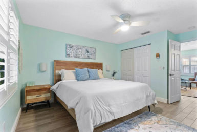 Coastal-style master bedroom with mint-green walls, wooden platform bed topped with white linens and blue accent pillows, mid-century nightstand, ceiling fan, plantation shutters, and sliding closet doors.