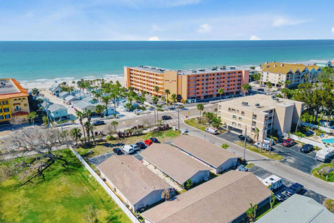 Aerial view of a sunny beachfront neighborhood with turquoise ocean and sandy beach, palm trees, a mid-rise orange hotel, rows of low-rise condos and parking lots along a coastal road — bright vacation resort scene.