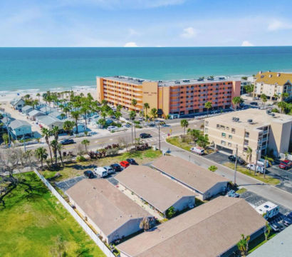 Aerial view of a sunny beachfront neighborhood with turquoise ocean and sandy beach, palm trees, a mid-rise orange hotel, rows of low-rise condos and parking lots along a coastal road — bright vacation resort scene.