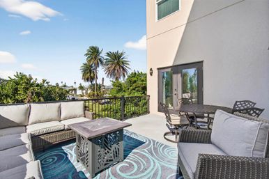 Sunny coastal balcony with wicker sectional and armchairs around a square coffee table on a turquoise swirled rug, outdoor dining set nearby, black railing and palm trees framing a marina view under a blue sky.