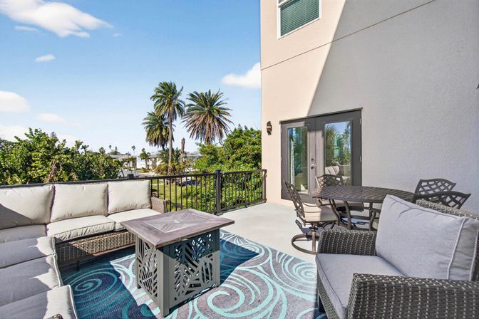 Sunny coastal balcony with wicker sectional and armchairs around a square coffee table on a turquoise swirled rug, outdoor dining set nearby, black railing and palm trees framing a marina view under a blue sky.