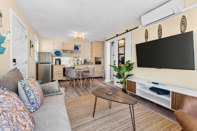 Bright open-plan living room and kitchenette in a coastal-style vacation rental — gray sofa with patterned pillows, mid-century wood coffee table, wall-mounted flat-screen TV, four-seat dining table, compact kitchen with stainless fridge and light wood cabinets, sliding barn door, indoor plant, and wall-mounted air conditioner.