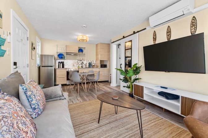 Bright open-plan living room and kitchenette in a coastal-style vacation rental — gray sofa with patterned pillows, mid-century wood coffee table, wall-mounted flat-screen TV, four-seat dining table, compact kitchen with stainless fridge and light wood cabinets, sliding barn door, indoor plant, and wall-mounted air conditioner.