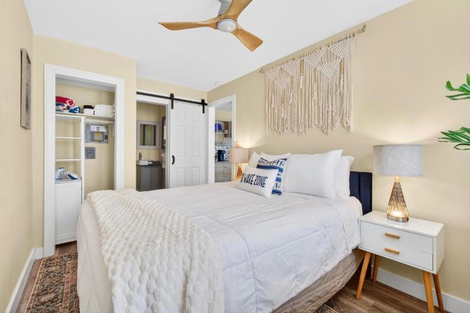 Bright coastal boho bedroom with plush white bedding, macramé wall hanging, wooden ceiling fan, sliding barn door to en-suite, mid-century nightstand and lamp