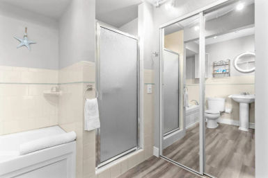 Bright coastal-style bathroom with frosted glass shower door, soaking tub with rolled towel, pedestal sink and toilet, mirrored sliding closet, starfish wall decor and wood-look flooring.