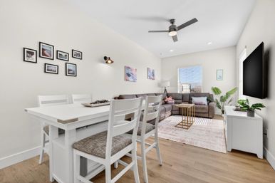 Bright modern open-concept apartment living room and dining area with white dining table and cushioned chairs, gray sectional sofa on a pale pink rug, wall-mounted TV, ceiling fan, hardwood floors, gallery wall and potted plants by a sunlit window.