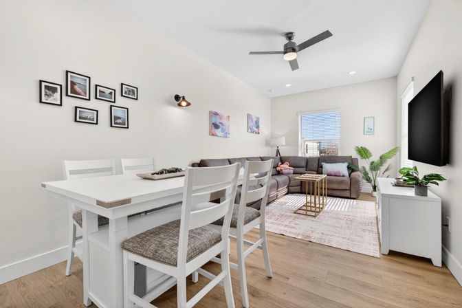 Bright modern open-concept apartment living room and dining area with white dining table and cushioned chairs, gray sectional sofa on a pale pink rug, wall-mounted TV, ceiling fan, hardwood floors, gallery wall and potted plants by a sunlit window.
