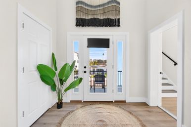 Bright coastal foyer with glass front door opening to a balcony and ocean view, potted tropical plant, round jute rug, woven wall hanging and staircase.