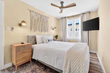 Cozy bright boho bedroom with queen bed dressed in white bedding, macramé wall hanging, wooden nightstands, brass sconces, ceiling fan, wall-mounted TV, and sunlit window.