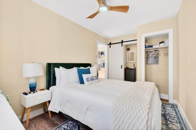 Sunny coastal-style bedroom with green velvet headboard, crisp white linens and blue accent pillows, wooden ceiling fan, bedside lamp on mid-century nightstand, patterned rug, and sliding barn-door closet.