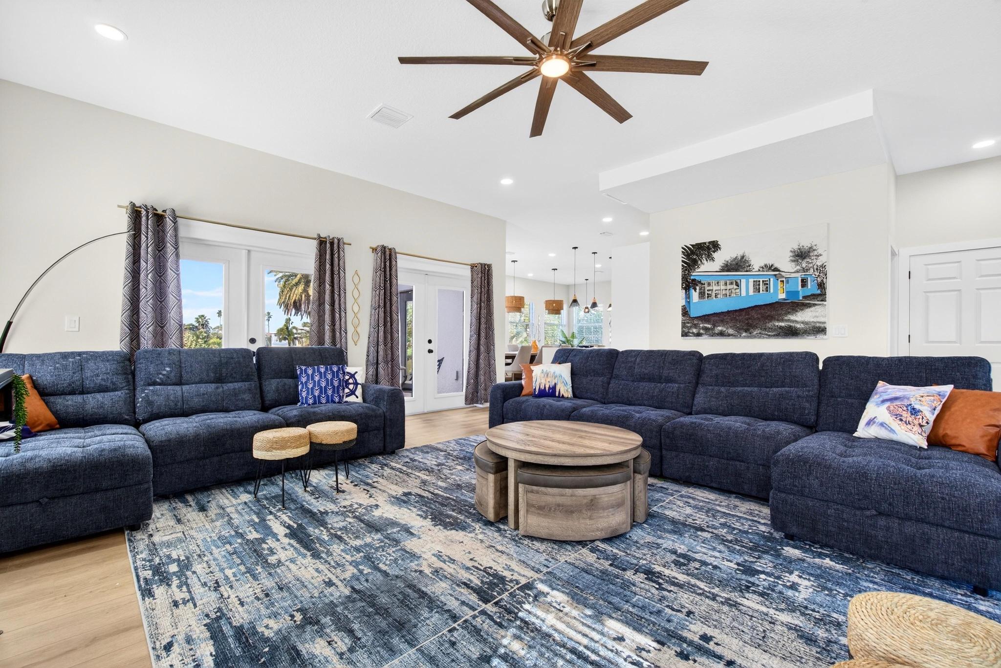 Bright open-plan living room with navy sectional sofas, round wooden nesting coffee tables, blue abstract area rug, large modern ceiling fan and patio doors showing palm trees — modern coastal style, sunlit and inviting.