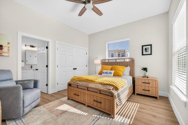 Sunlit coastal-style bedroom with a wooden storage bed and matching nightstand, mustard-yellow throw and pillows, gray armchair, ceiling fan, hardwood floors, window blinds, and an adjacent bathroom and double-door closet.