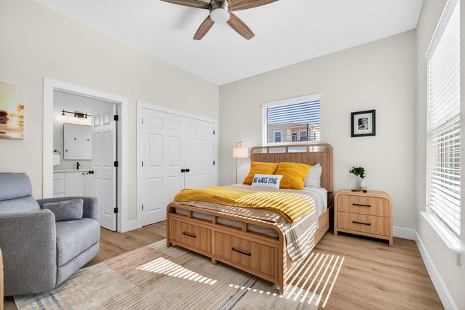 Sunlit coastal-style bedroom with a wooden storage bed and matching nightstand, mustard-yellow throw and pillows, gray armchair, ceiling fan, hardwood floors, window blinds, and an adjacent bathroom and double-door closet.