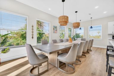 Bright sunlit dining room with a long wood table and ten gray upholstered chairs, rattan pendant lights, large windows and tropical garden views
